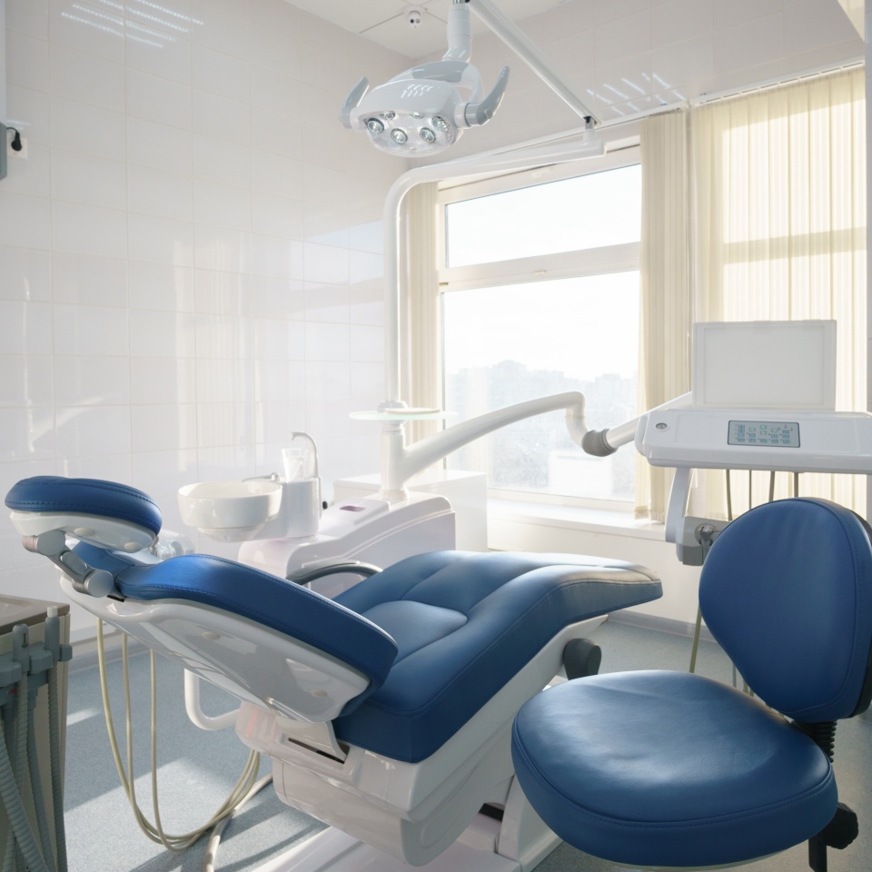 A blue dental chair, overhead light, and control unit in a clinical exam room.